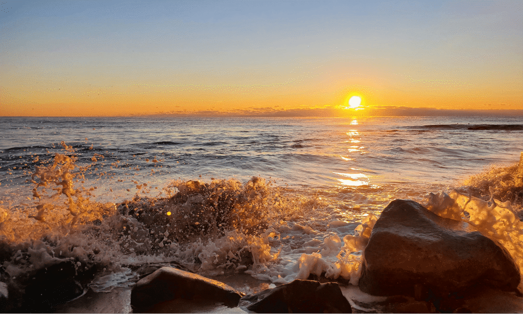 Waves crashing over dark rocks on a rocky coastline at sunset, with the sun low on the horizon casting a golden reflection across the water