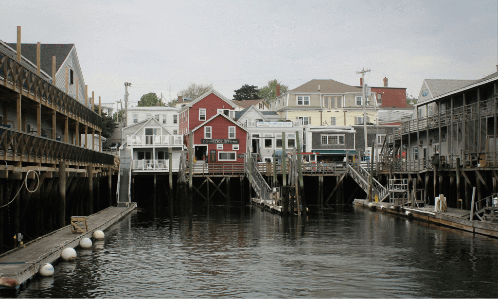 A harbor lined with wooden docks and pilings, with weathered clapboard buildings including a red Country Store visible along the waterfront on an overcast day
