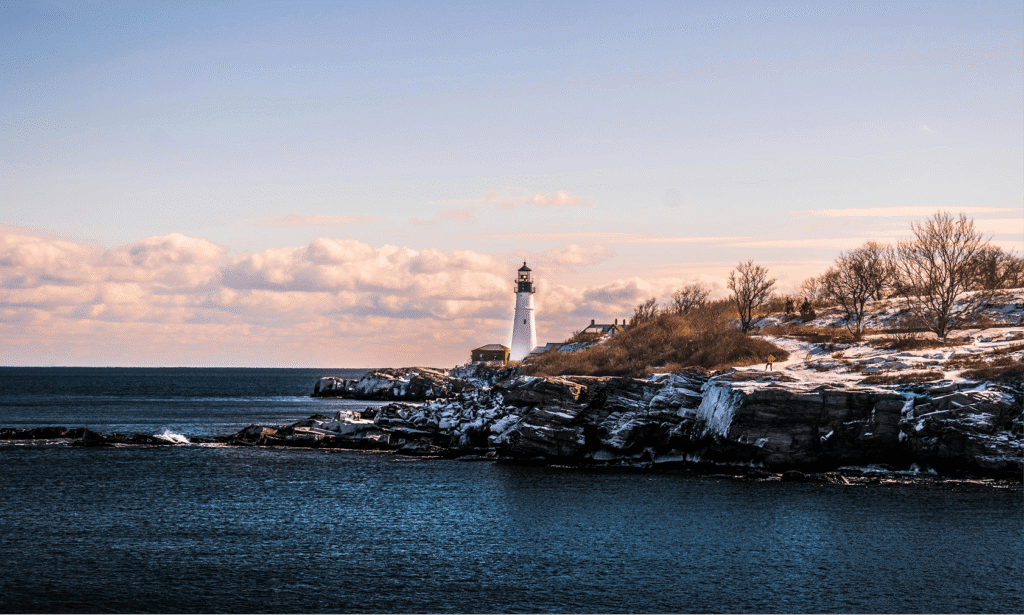 A white lighthouse on a snow-dusted rocky coastline at golden hour, surrounded by bare winter trees and jagged ledge, with calm dark water in the foreground