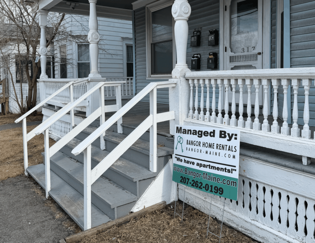 A gray clapboard multi-unit rental house with a white wrap-around porch, front steps, and a Bangor Home Rentals yard sign in the foreground
