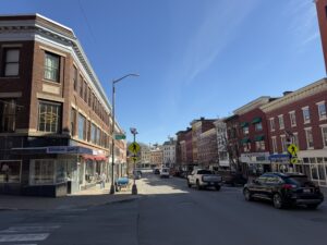 Historic brick storefronts along Main Street in downtown Bangor, Maine, on a clear sunny day with parked cars and a pedestrian.