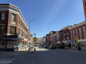 Historic brick storefronts along Main Street in downtown Bangor, Maine, at the Cross Street intersection on a clear spring day.
