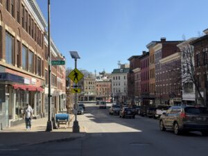 Main Street at Cross Street in downtown Bangor, Maine, with historic brick storefronts, parked cars, and a pedestrian crosswalk sign on a sunny day
