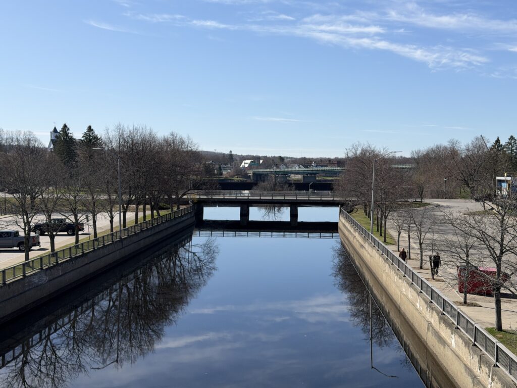 Kenduskeag Stream flowing between concrete flood walls in downtown Bangor, Maine, with a bridge and church steeple on a clear spring day.