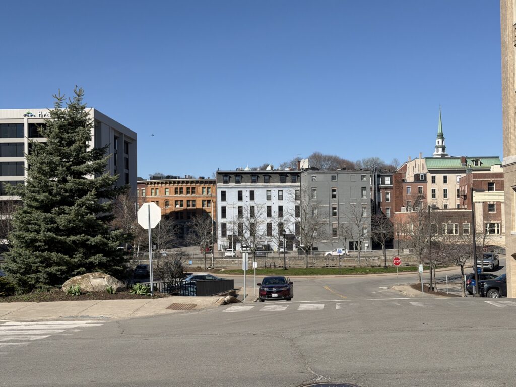 Historic brick and painted rowhouse buildings with a church steeple in downtown Bangor, Maine, on a clear spring day.