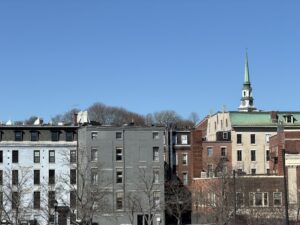 Historic downtown Bangor, Maine skyline with brick and clapboard apartment buildings and a green copper church steeple against a clear blue sky.