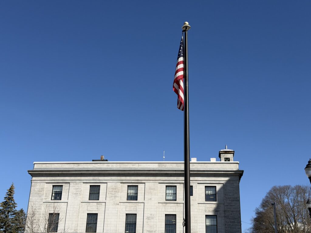 American flag on a tall flagpole in front of a historic granite government building under a clear blue sky