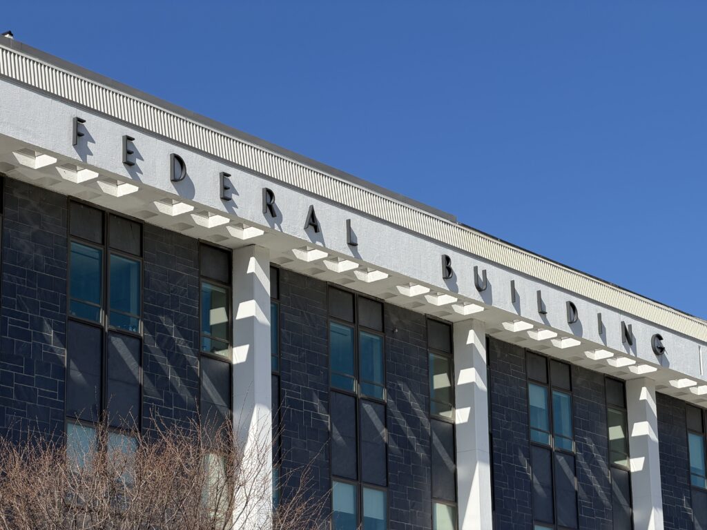 Low-angle view of the Margaret Chase Smith Federal Building in Bangor, Maine, with raised metal "Federal Building" lettering, dark stone panels, and a white cornice against a blue sky.