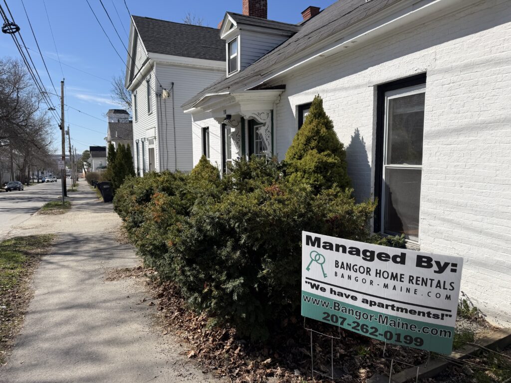White painted brick rental home with an ornate front entry and evergreen shrubs, with a "Managed By: Bangor Home Rentals" sign in the yard beside a sidewalk in Bangor, Maine.