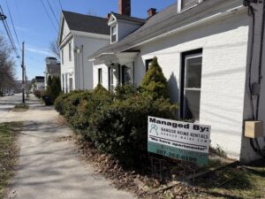 Managed By Bangor Home Rentals sign in front of a white multi-unit apartment house on a residential street in Bangor, Maine
