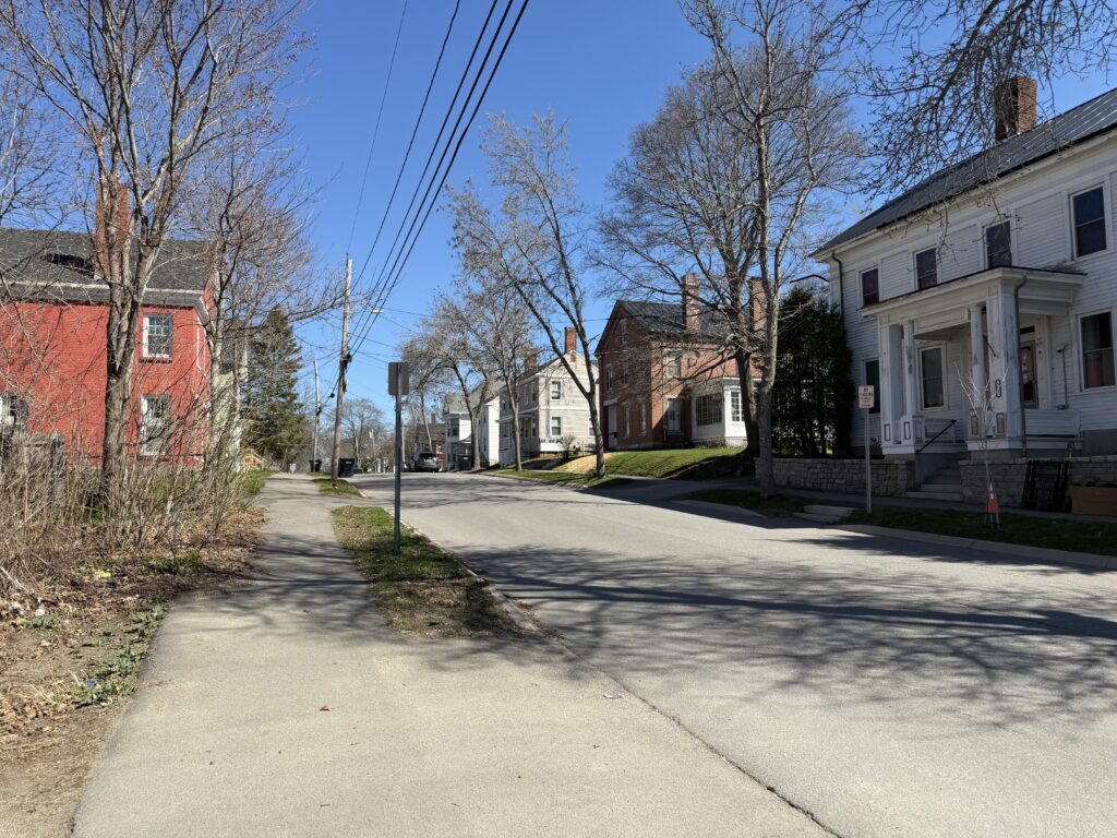 Quiet residential street in Bangor, Maine, lined with historic clapboard and brick homes on a clear spring day