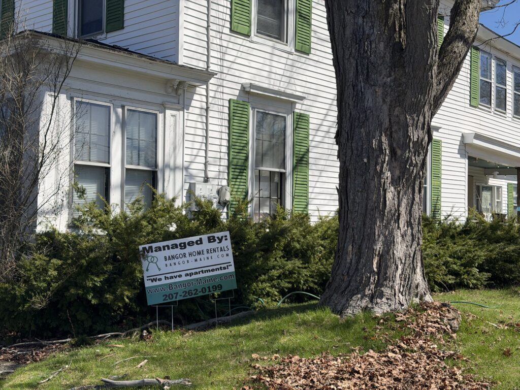White two-story multi-unit house with green shutters and a bay window, with a Bangor Home Rentals "Managed By" sign in the front yard
