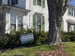 White two-story multi-unit house with green shutters and a bay window, with a Bangor Home Rentals "Managed By" sign in the front yard