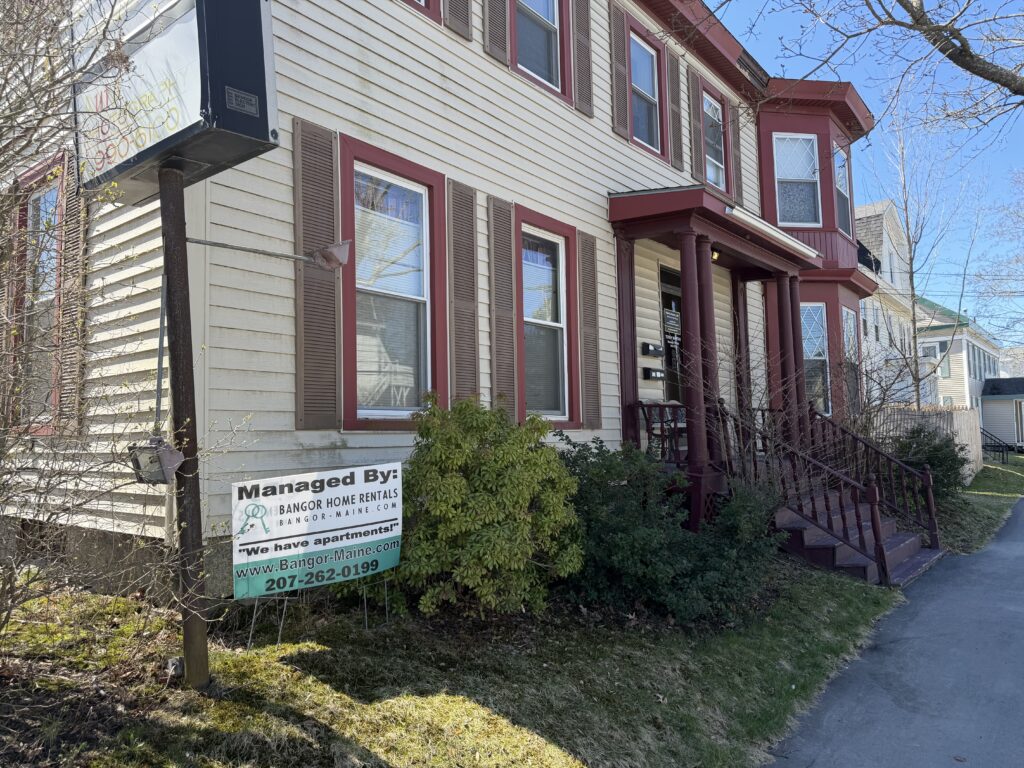 Two-story apartment building with cream siding and burgundy trim in Bangor, Maine, with a Bangor Home Rentals sign in the yard.