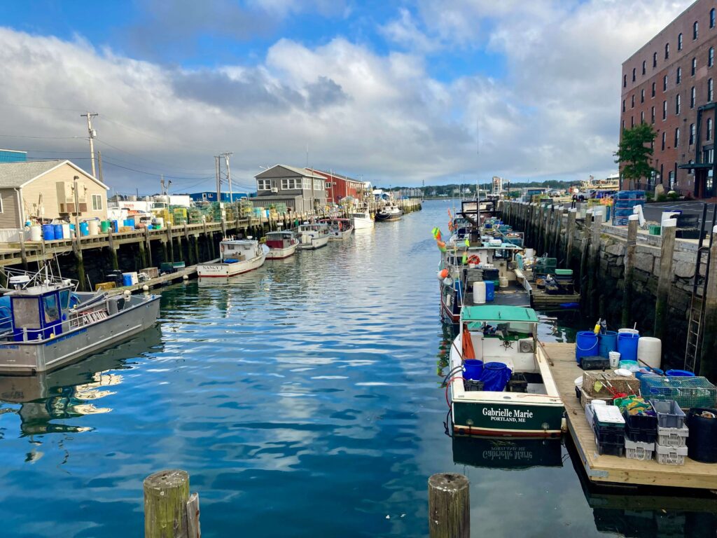 Portland, Maine working waterfront with lobster boats moored along Commercial Street docks, lobster traps stacked on piers, and the brick Old Port district visible at right. Foreground vessel reads "Gabrielle Marie, Portland, ME."