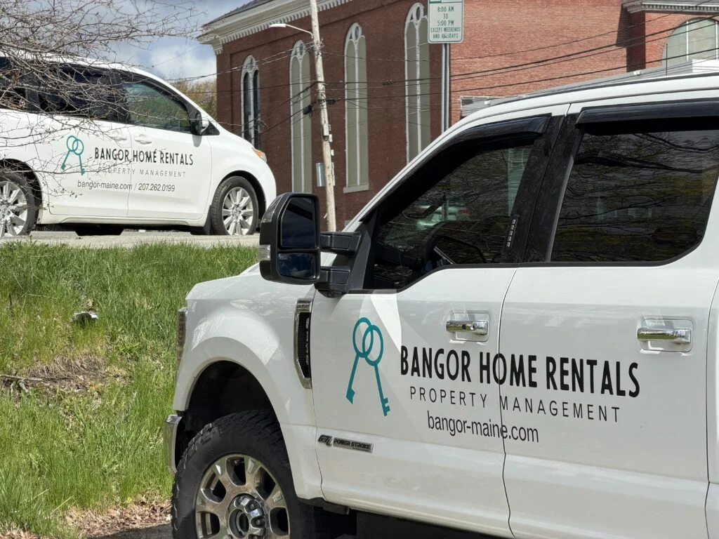 Two white Bangor Home Rentals property management vehicles with teal key logo parked near a brick building in Bangor, Maine